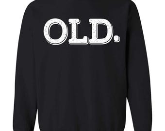 May include: A black long-sleeve crewneck sweatshirt featuring the word OLD. printed in large, white, distressed-style capital letters across the chest. The text includes a period at the end and has a subtle textured appearance on the lettering.