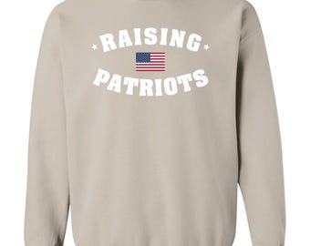 May include: A beige crewneck sweatshirt featuring the white text RAISING PATRIOTS arched above and below a small graphic of the United States flag. The design includes two small white stars on either side of the word RAISING.