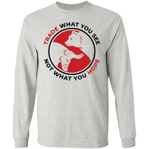 May include: A light gray long-sleeve shirt featuring a circular graphic with a red background. Inside the circle are white silhouettes of a bear and a bull. The text reads TRADE WHAT YOU SEE NOT WHAT YOU HOPE in black and red capital letters.