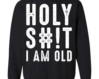 May include: A black long-sleeve sweatshirt featuring white distressed-style text on the front. The graphic displays the words HOLY S#!T I AM OLD in a bold, sans-serif font. The design is centered on the chest of the garment, which has a crew neckline.