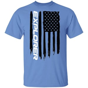 May include: A light blue short-sleeve t-shirt featuring a black graphic of a distressed American flag oriented vertically. The word EXPLORER is printed in large white capital letters vertically along the left side of the flag graphic.