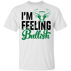 May include: A white short-sleeved t-shirt featuring a graphic print with the text I'M FEELING Bullish in black and green. The design includes a green bull head illustration and a green upward-trending line graph integrated into the text.