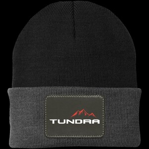 May include: A two-tone knit beanie featuring a black crown and a dark gray folded cuff. Centered on the cuff is a rectangular patch with a dark gray background, displaying the white text TUNDRA below a stylized red mountain graphic.