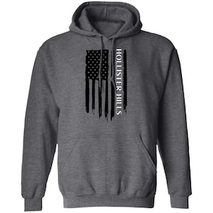 May include: A dark heather gray hooded sweatshirt featuring a black distressed American flag graphic on the chest. The text HOLLISTER HILLS is printed vertically in white capital letters alongside the flag design. It has long sleeves and a front pocket.