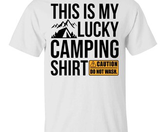 May include: A white short-sleeved t-shirt featuring black text that reads THIS IS MY LUCKY CAMPING SHIRT with a graphic of mountains and a tent. Below the text is an orange rectangular caution sign with black text reading CAUTION DO NOT WASH.