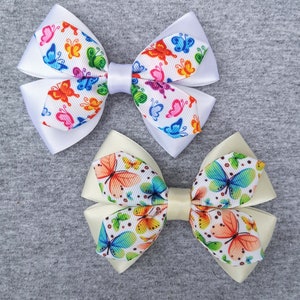 May include: Two white satin hair bows with butterfly patterns. One bow has a white background with colourful butterflies, the other has a cream background with blue, orange, and green butterflies.