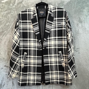 May include: A black and white plaid blazer with a single-button closure and two front pockets. The jacket is hanging on a black hanger against a gray background. The blazer has a classic, tailored design.