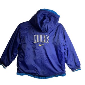 May include: A blue Nike jacket with a hood. The jacket has a white and yellow Nike logo on the back. The jacket has blue and white striped cuffs and a blue and white striped hem.