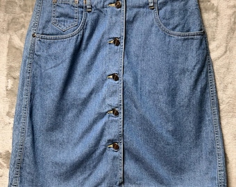 May include: A light blue denim skirt with a button-up front closure. The skirt has two front pockets and a small pocket on the right side. The skirt is hanging on a white hanger.