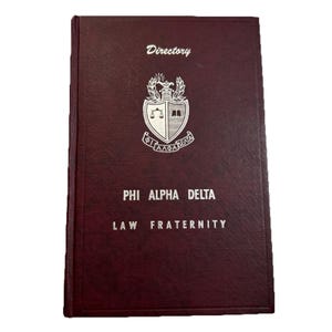 May include: A maroon directory with the text "Directory" at the top. The cover features a white emblem with scales, a book, and the Greek letters "ΦΑΔ". Below, it reads "PHI ALPHA DELTA LAW FRATERNITY".