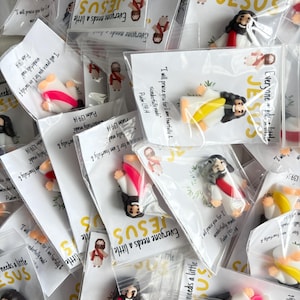 May include: Small Jesus figurines in clear plastic bags, scattered across a surface. Each bag has a printed card with the text "Everyone needs a little JESUS" and a Bible verse. The figurines depict Jesus in various poses, with colorful robes.
