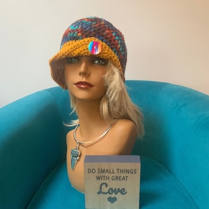 May include: A colorful crocheted hat with a yellow brim and a rainbow button. The hat is on a mannequin head with a silver necklace and a turquoise pendant. The mannequin is sitting on a teal blue chair. A wooden sign with the text "Do small things with great love" is in front of the mannequin.