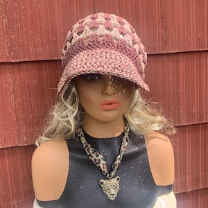 May include: A crochet hat in shades of pink and beige with a brim. The hat is displayed on a mannequin with a leopard print necklace. The background is a red wooden wall.