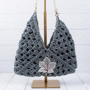 May include: A gray crocheted handbag with a leaf applique. The bag has a unique open weave design and a pearl chain strap. The bag is displayed on a gold stand against a white wooden background. The leaf applique is gray and brown.