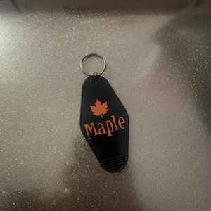 May include: Black motel key chain with a silver key ring. The key chain is shaped like a motel sign with the word "Maple" and a stylized orange maple leaf.