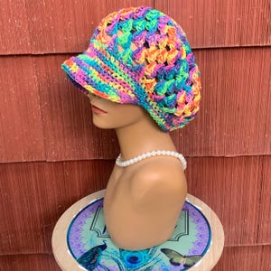 May include: A vibrant, multi-colored crochet newsboy cap featuring a variegated yarn pattern in shades of neon pink, bright yellow, electric blue, and lime green. The hat has a structured brim and a textured, open-stitch design throughout.