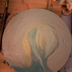 May include: A round canvas with a textured surface, painted with a light blue and yellow abstract design. The design resembles a person's head and shoulders, with the hair painted in yellow and the shoulders in blue.
