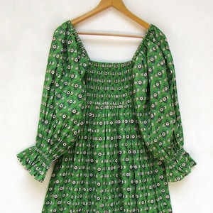 Könnte beinhalten: Ein grünes Kleid mit einem sich wiederholenden Blumenmuster in Weiß und Marineblau. Das Kleid hat lange Ärmel mit Rüschenbündchen, einen quadratischen Ausschnitt und ein gerafftes Oberteil. Das Kleid hängt an einem Holzkleiderbügel.