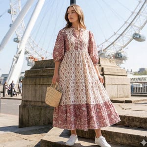 May include: A patterned maxi dress in mauve, cream, and white, with three-quarter sleeves and a V-neck. The dress is tiered and worn with white trainers. A small straw tote bag is carried. The London Eye is visible in the background.