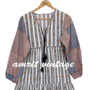 Op de afbeelding: Een lange, gelaagde jurk met een V-hals en lange mouwen. De jurk heeft een mix van patronen, waaronder wit met bruine en zwarte pijldessins, en delen van rood en blauw geruite stof. De tekst "amrit vintage" is zichtbaar.