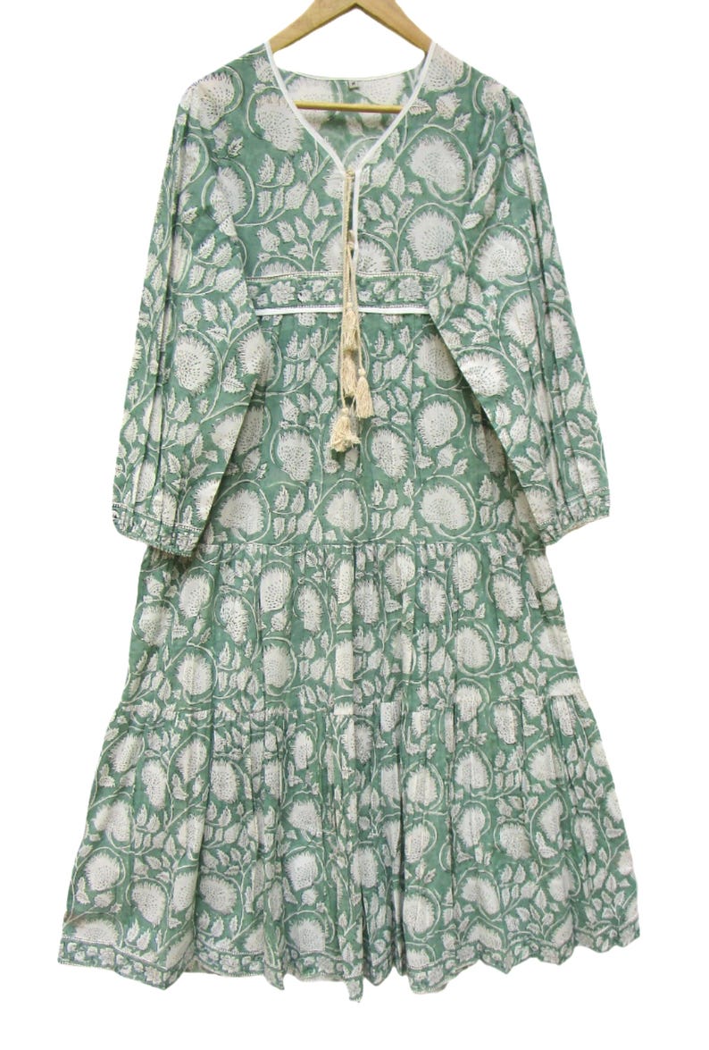 May include: A long, flowing dress in a soft green colour, decorated with a white floral pattern. The dress has three-quarter sleeves, a V-neckline with a tassel tie, and a tiered skirt. The fabric appears lightweight and suitable for warm weather.