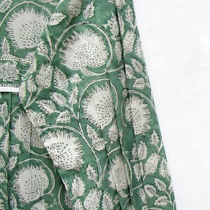 May include: A green dress with a white floral pattern. The dress has a V-neck and long sleeves. The fabric is printed with large white flowers and leaves on a green backdrop. The sleeves have gathered cuffs. The dress has a ruffled hem.