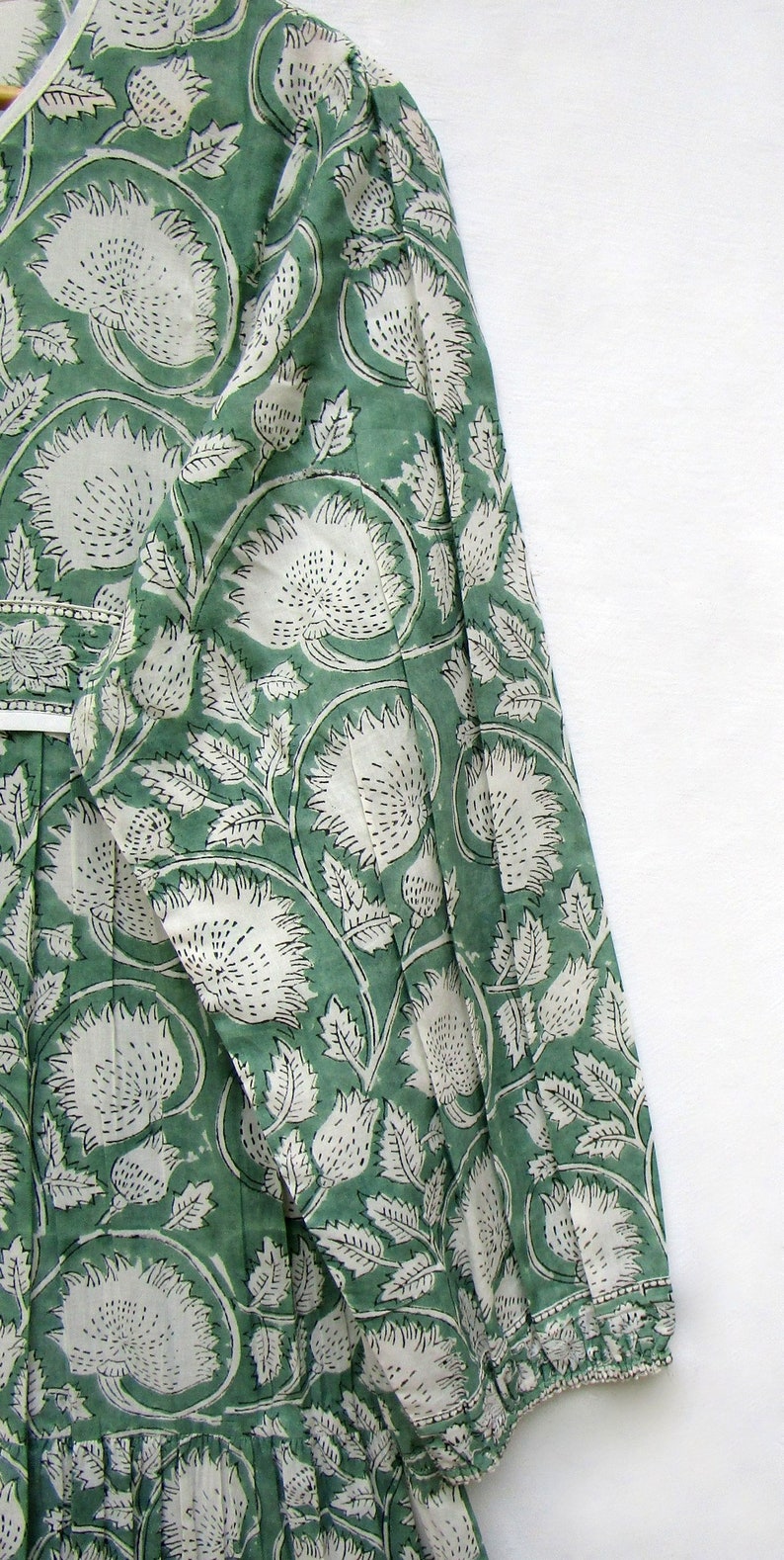 May include: A green dress with a white floral pattern. The dress has a V-neck and long sleeves. The fabric is printed with large white flowers and leaves on a green backdrop. The sleeves have gathered cuffs. The dress has a ruffled hem.