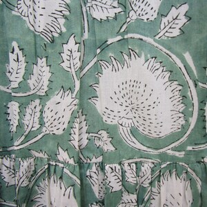 May include: Close-up of a fabric with a repeating floral pattern. The design features large white flowers and leaves with black outlines against a muted green background. The fabric has a textured appearance.