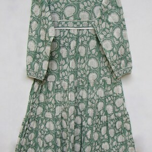 May include: A long, green dress with a white floral pattern. The dress has long sleeves, a tiered skirt, and a white trim around the neckline and waist. The dress is hanging on a wooden hanger.