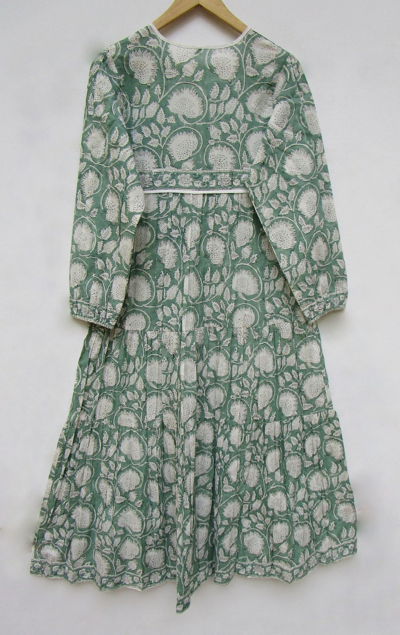 May include: A long, green dress with a white floral pattern. The dress has long sleeves, a tiered skirt, and a white trim around the neckline and waist. The dress is hanging on a wooden hanger.