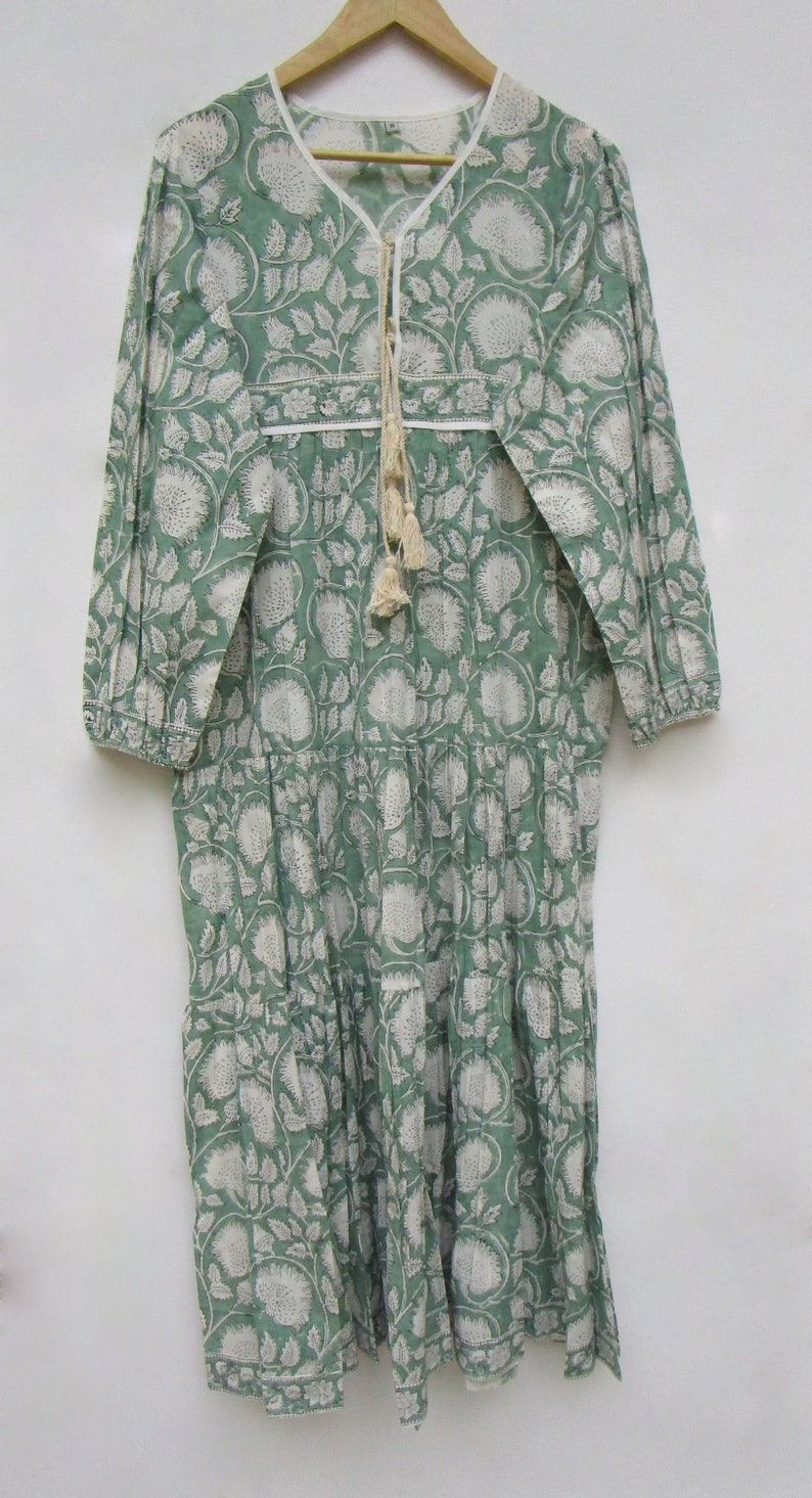 May include: A long, green dress with a white floral print. It has long sleeves, a V-neck with a tassel tie, and a tiered skirt. The dress is made of a lightweight fabric, ideal for warmer weather. The measurements are in centimetres.