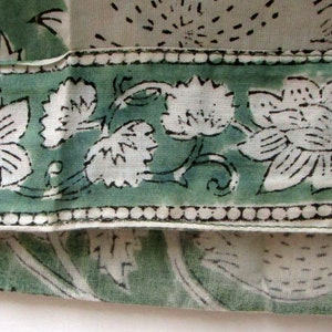 May include: Close-up of a fabric with a floral pattern. The design features large white flowers with black outlines and green leaves on a light green border. The background is a light cream colour.