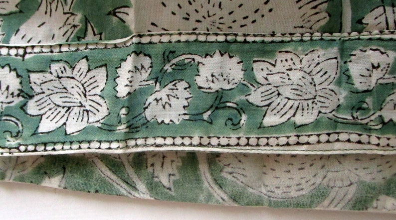 May include: Close-up of a fabric with a floral pattern. The design features large white flowers with black outlines and green leaves on a light green border. The background is a light cream colour.