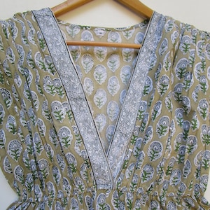 May include: A beige cotton dress with a V-neckline and a floral print. The dress has a white and gray trim around the neckline. The dress is on a wooden hanger. The size tag says "M".