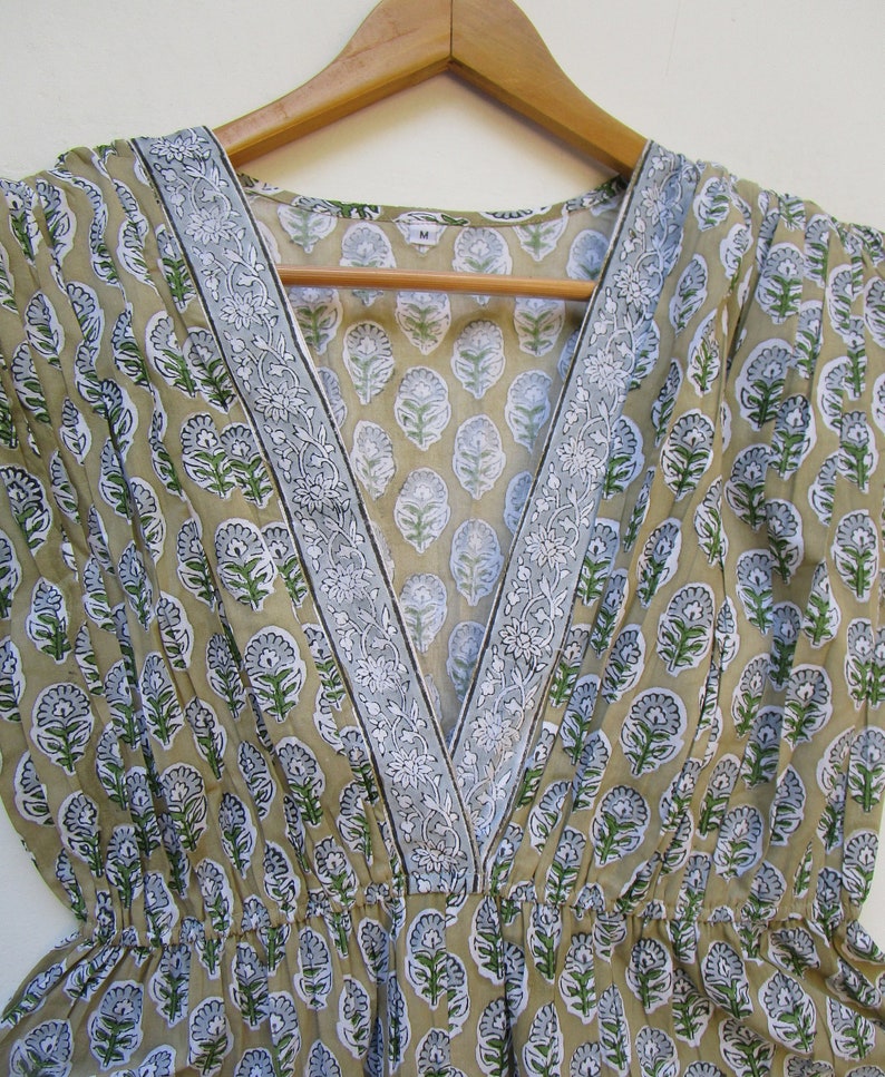 May include: A beige cotton dress with a V-neckline and a floral print. The dress has a white and gray trim around the neckline. The dress is on a wooden hanger. The size tag says "M".