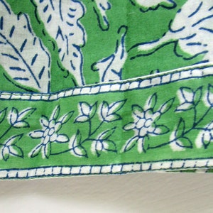 May include: Green fabric with a white floral pattern and a blue trim. The fabric has a white border with a repeating pattern of small white flowers.