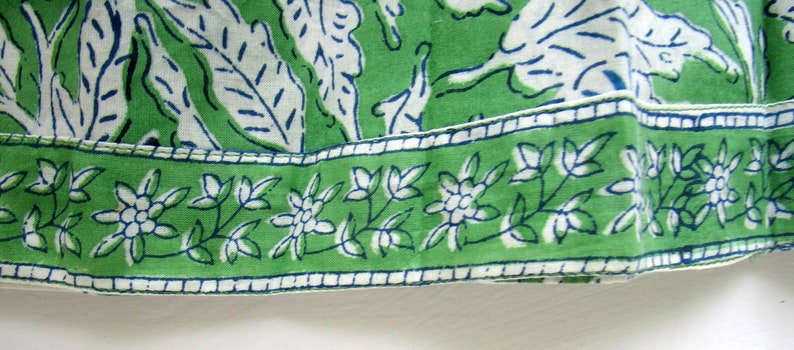 May include: Green fabric with a white floral pattern and a blue trim. The fabric has a white border with a repeating pattern of small white flowers.