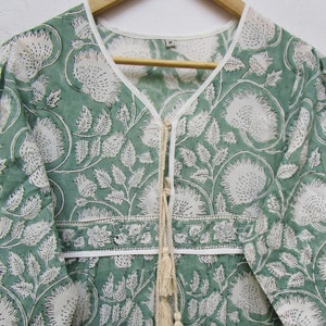 May include: A green and white floral print dress with a V-neck and long sleeves. The dress has a white trim along the neckline and a decorative tie with tassels. The fabric features a repeating pattern of large flowers and leaves.