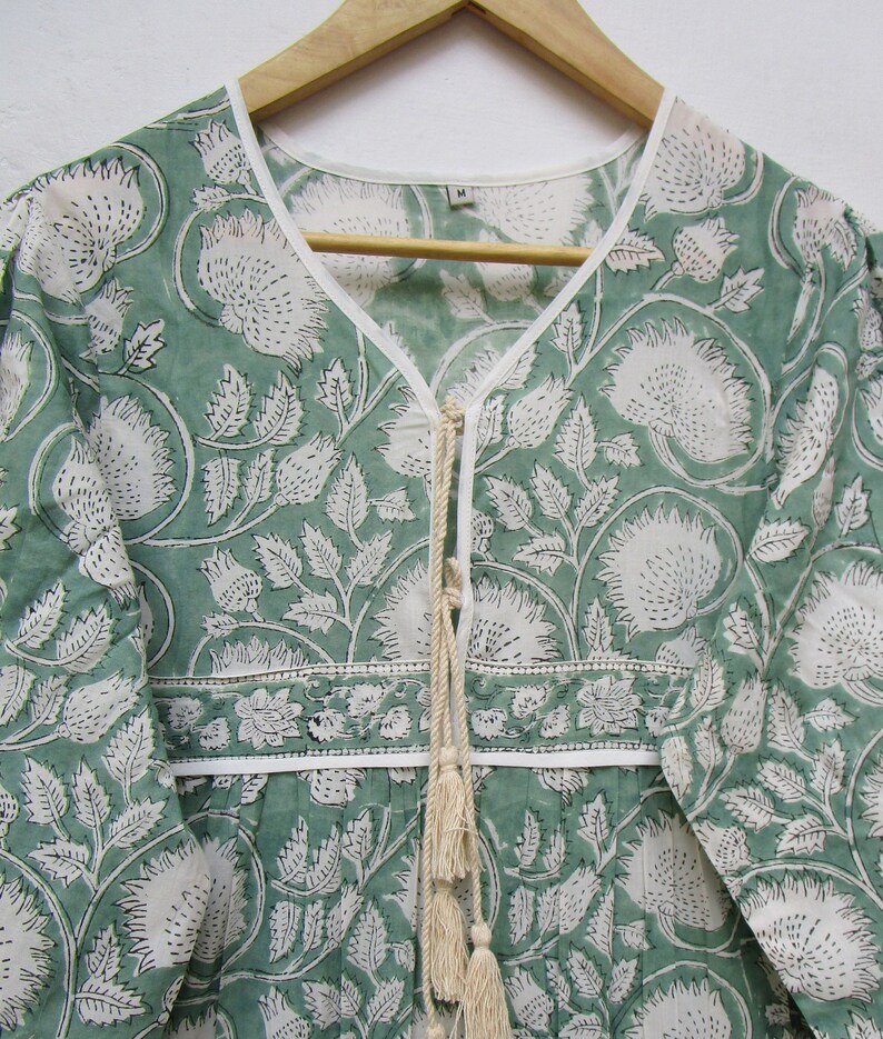 May include: A green and white floral print dress with a V-neck and long sleeves. The dress has a white trim along the neckline and a decorative tie with tassels. The fabric features a repeating pattern of large flowers and leaves.