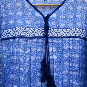 May include: A blue cotton tunic top with a white floral print and a navy blue tassel tie at the neckline. The top has a white and blue geometric border.