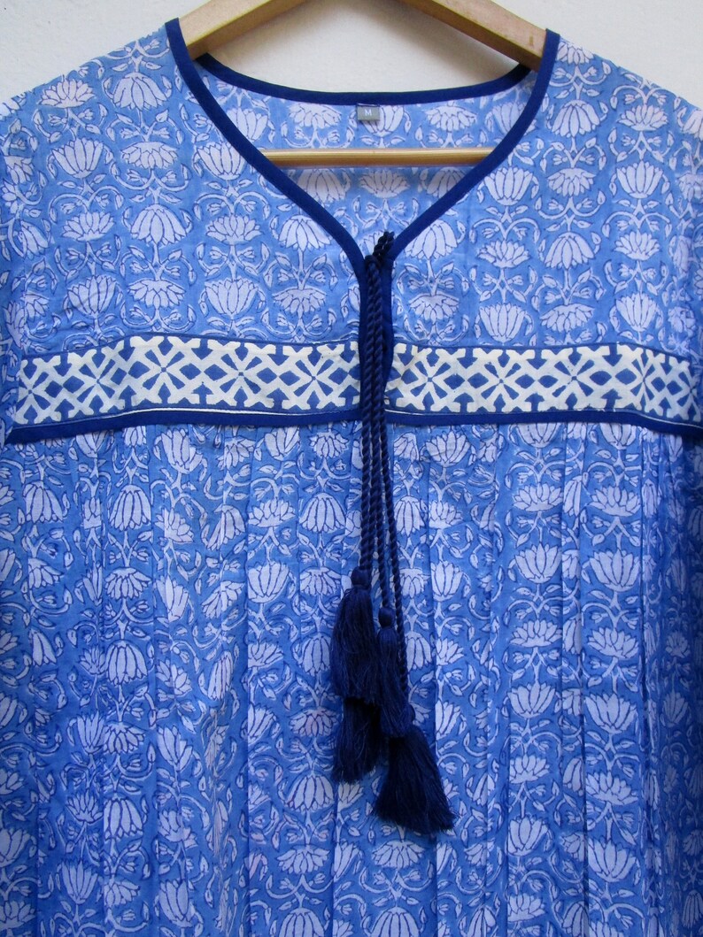 May include: A blue cotton tunic top with a white floral print and a navy blue tassel tie at the neckline. The top has a white and blue geometric border.