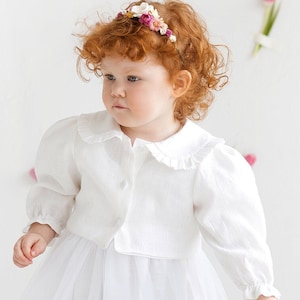 May include: A young girl with red curly hair wears a white dress with a ruffled collar and a flower crown. She is standing in front of a white background with pink tulips scattered around her. She is holding a bucket of pink and purple tulips.