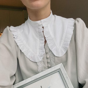 Może przedstawiać: Szara bluzka z białym falbaniastym kołnierzykiem i koronkową lamówką. Bluzka ma rząd guzików z przodu. Osoba trzyma oprawiony obraz budynku z wieżą.