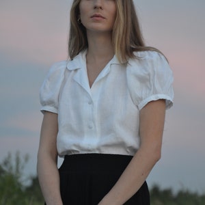 May include: A woman wearing a white linen blouse with puffed sleeves and a black skirt. She is standing in front of a blurred background of greenery and a cloudy sky.