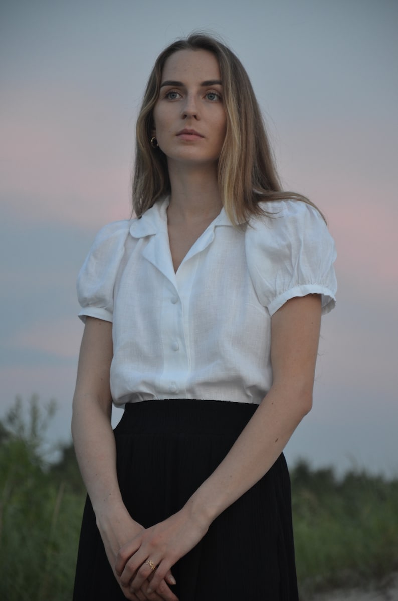 May include: A woman wearing a white linen blouse with puffed sleeves and a black skirt. She is standing in front of a blurred background of greenery and a cloudy sky.