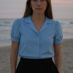 May include: A woman wearing a light blue collared shirt with puffed sleeves and black pants. She is standing on a sandy beach with the ocean in the background.