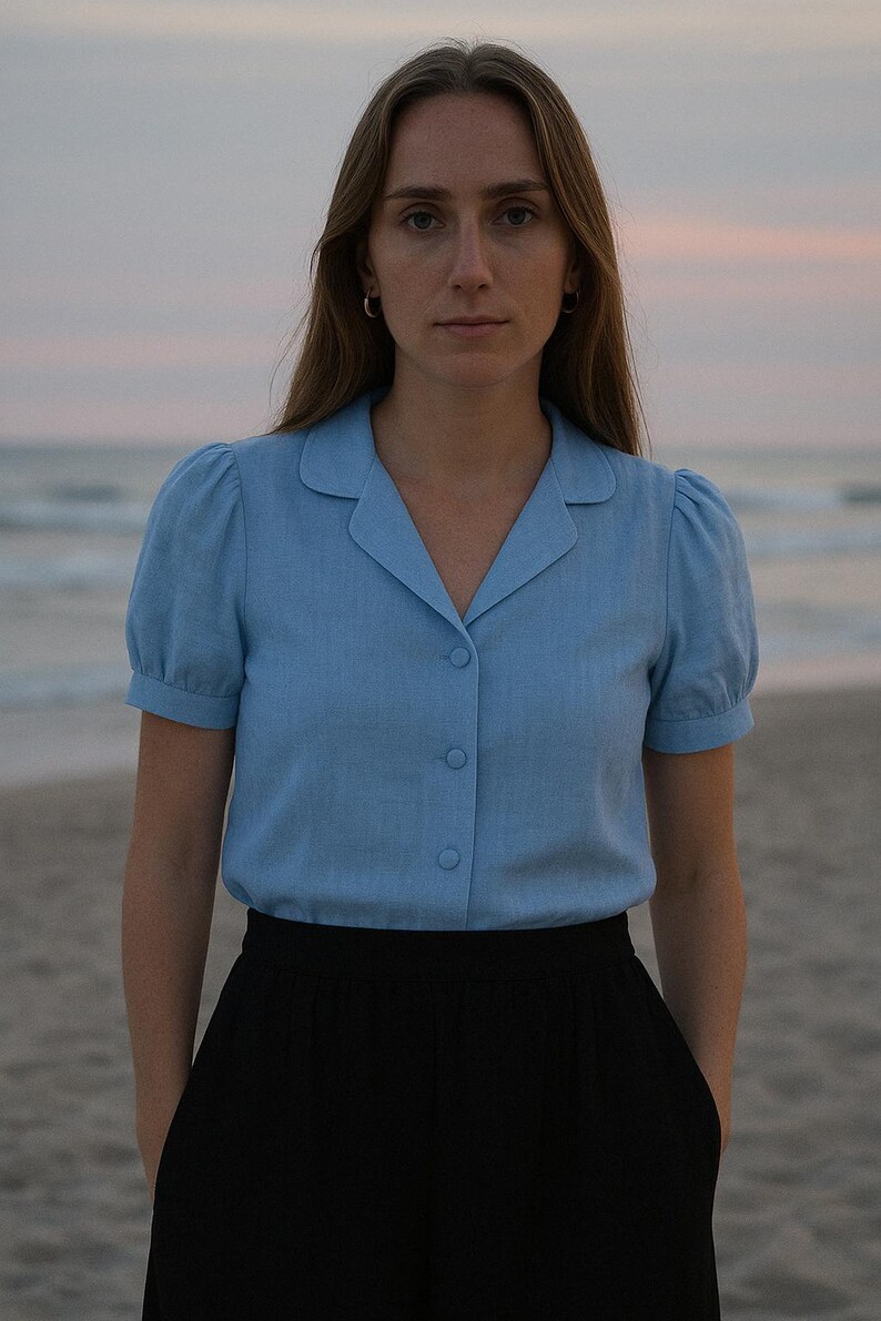 May include: A woman wearing a light blue collared shirt with puffed sleeves and black pants. She is standing on a sandy beach with the ocean in the background.