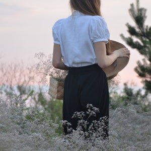 May include: A woman in a white blouse and black skirt stands in a field of white flowers. She is holding a straw hat in her right hand.