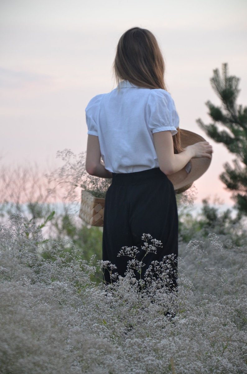 May include: A woman in a white blouse and black skirt stands in a field of white flowers. She is holding a straw hat in her right hand.