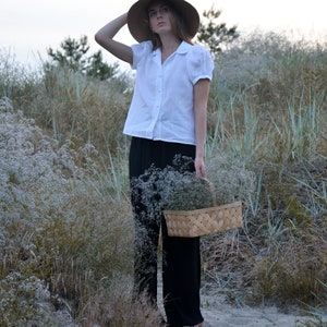 May include: A woman wearing a white linen shirt and black pants holds a woven basket filled with white flowers. She is standing in a field of tall grass and wildflowers.
